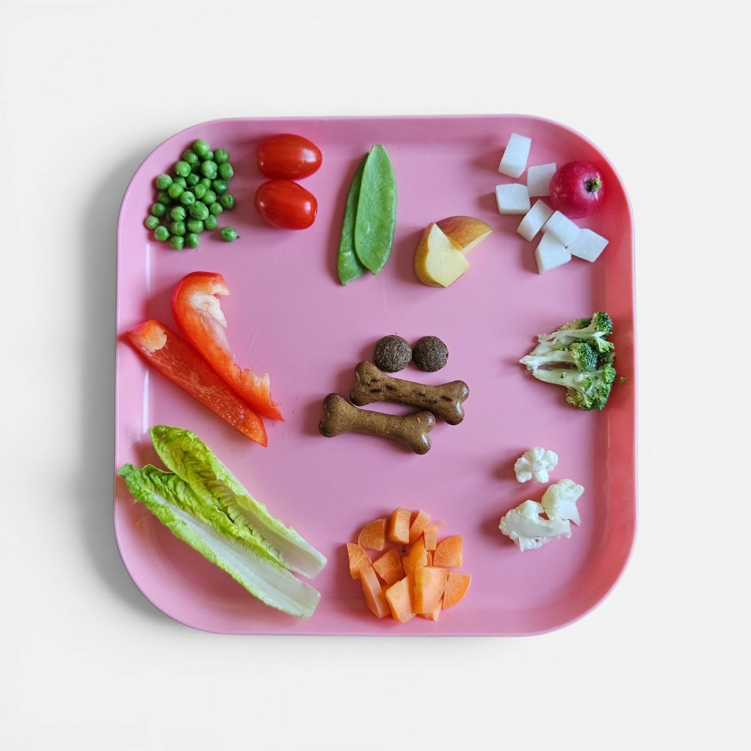 A pink tray laid with safe foods for a snuffle mat — peas, cherry tomatoes, mangetout, apple, cucumber, red pepper, romaine lettuce, carrot, broccoli, cauliflower, and bone-shaped treats